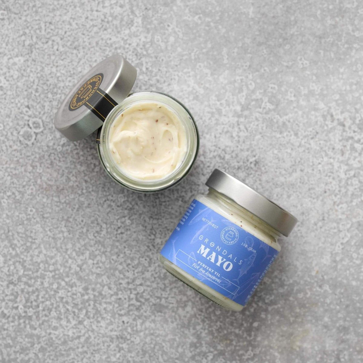 To glas Grøndals Mayonnaise – perfekt til fisk fra Grøndals, det ene åbent og afslører cremet mayonnaise med frilandsæg og det andet lukket med en blå etiket, står på en lysegrå tekstureret overflade – ideelt til servering med fiskekonserves.