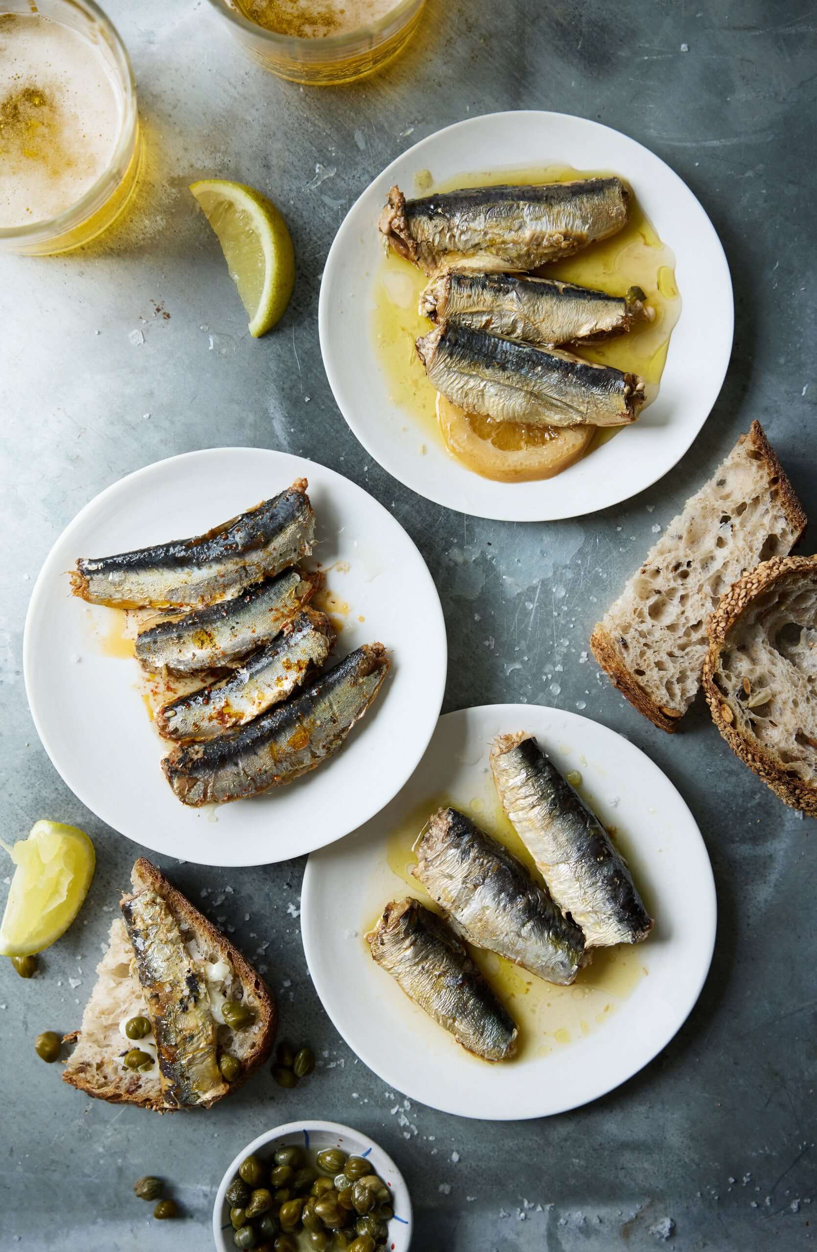 Tre hvide tallerkener med Jean de Luz Sardin-trio er arrangeret på en grå flade og serveres med rustikt brød, citronbåde, kapers og to glas let øl.