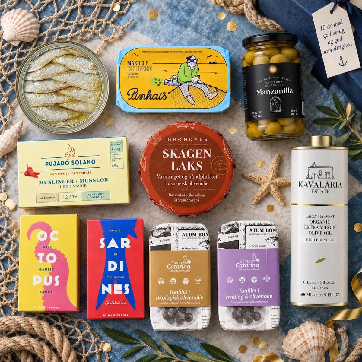 Et »flat lay«-billede med Grøndals’ 10-års jubilæumskasse, elegant anrettet med gourmetkonserves, et glas oliven, en flaske olivenolie og en lille skål med fisk – alt sammen anbragt på en lys stenoverflade, pyntet med reb og en papirlap, der tilbyder rabat.