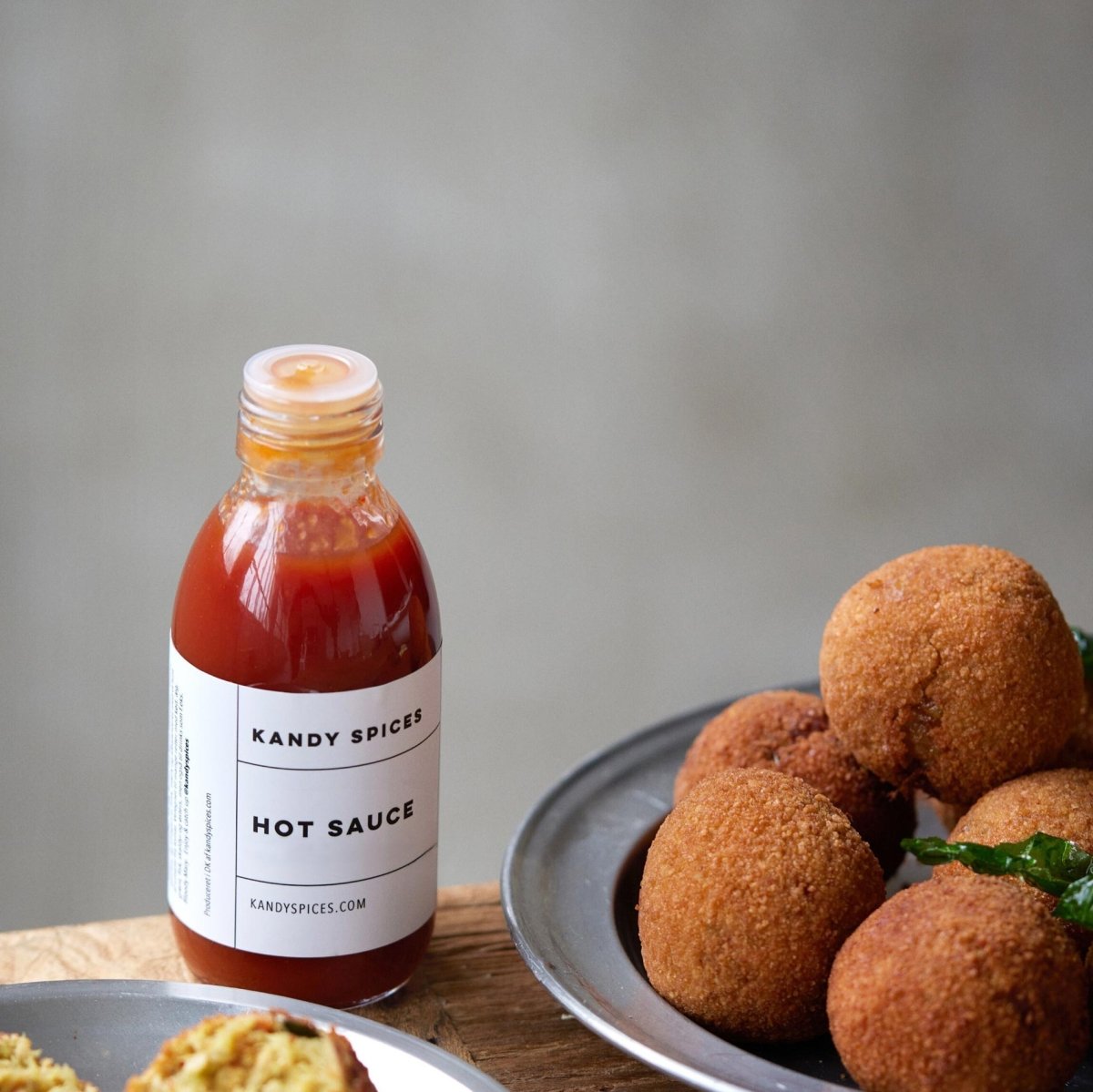 En flaske Kandy Spices HOT SAUCE fra Kandy Spices står på en træplade ved siden af en metaltallerken med gyldenbrune, stegte snacks på en almindelig, sløret baggrund.
