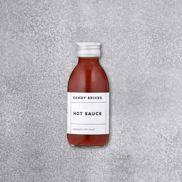En lille glasflaske med Kandy Spices HOT SAUCE med hvid etiket og sølvfarvet låg står på en grå, struktureret overflade. På etiketten står der "Habanero chilli sauce" under Kandy Spices' hovedbranding.