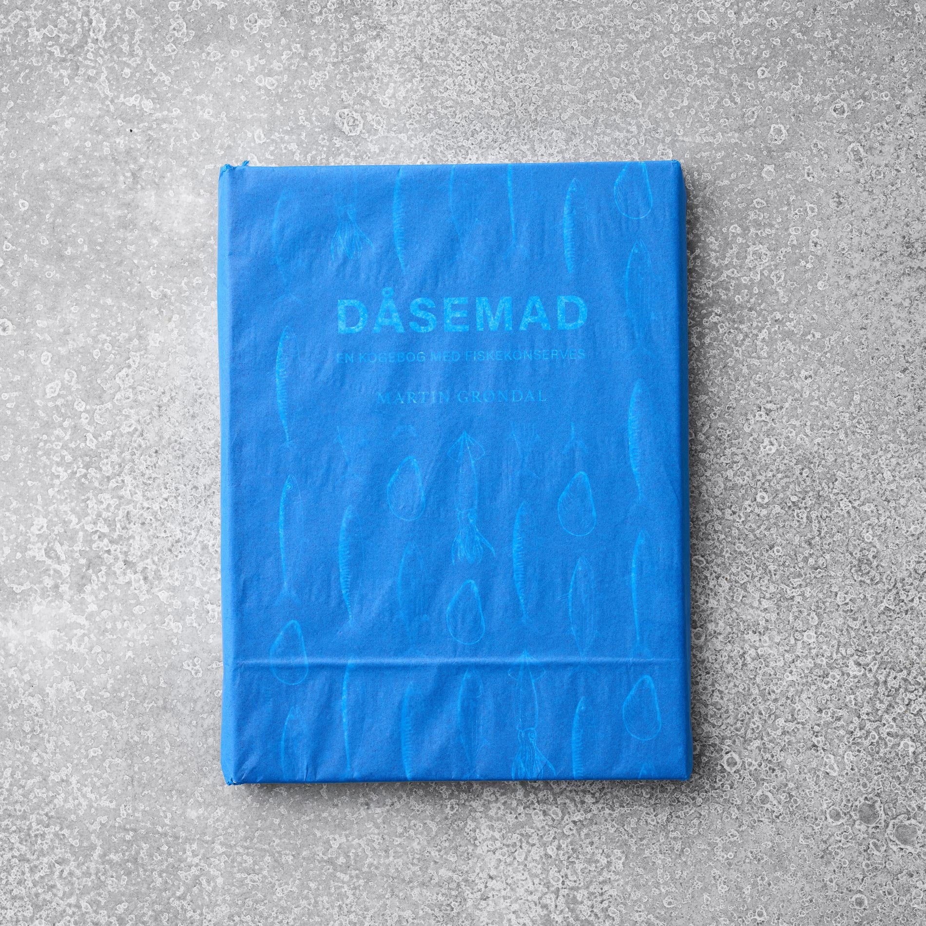 Den blå Kogebogen DÅSEMAD fra Grøndals hviler på en tekstureret grå overflade, hvis fiskeskælsmønster antyder kreative opskrifter på dåsefisk.