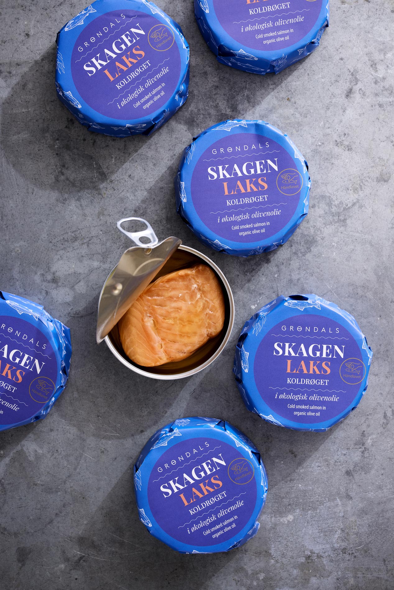 En dåse Koldrøget laks i økologisk olivenolie fra Grøndals står åben på en grå overflade, omgivet af fem uåbnede dåser af det samme produkt.