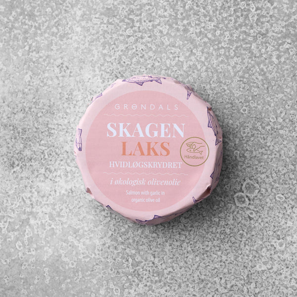 En rund dåse Grøndals Hvidløgskrydret laks i økologisk olivenolie med lyserød etikette hviler på en grå tekstureret overflade. Etiketten viser dansk og engelsk tekst om den hvidløgskrydrede laks i økologisk olivenolie.