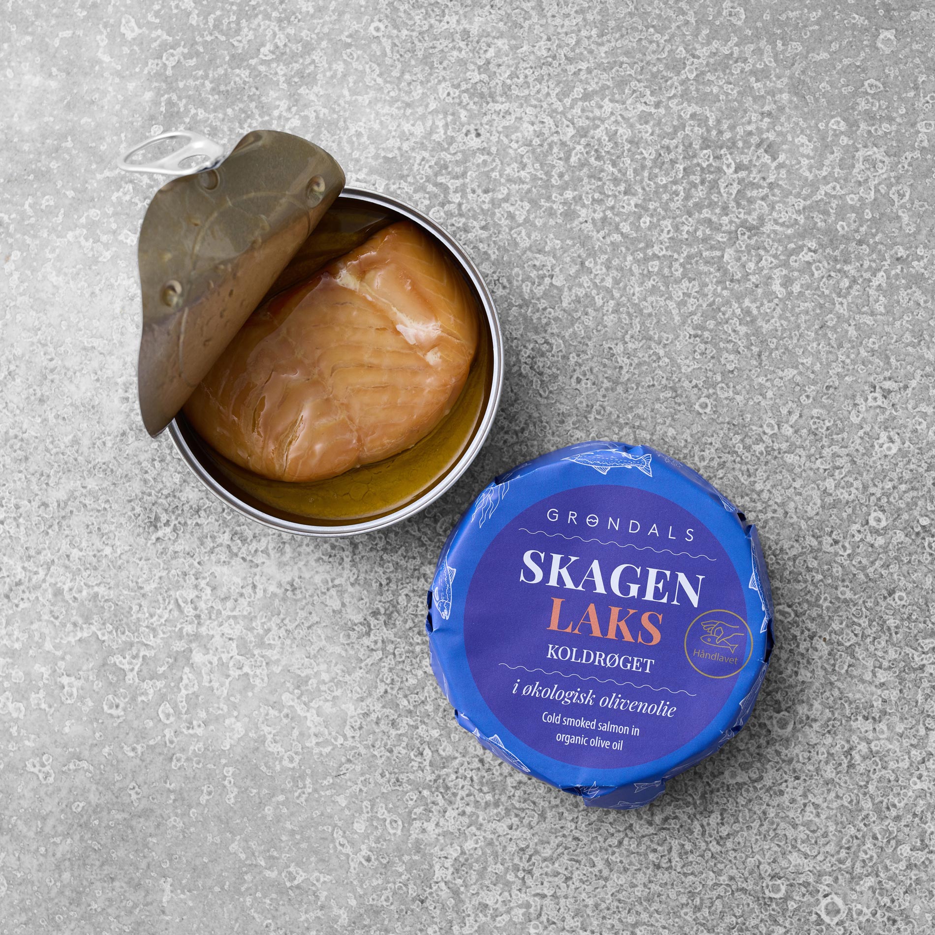 En åbnet dåse Grøndals Koldrøget laks i økologisk olivenolie ligger på en grå overflade. Det blå låg, mærket med mærke og produktnavn, ligger ved siden af dåsen.