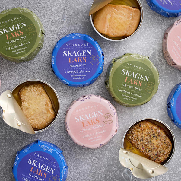 Grøndals lakse-trio runde dåser, nogle åbnet for at vise koldrøget dansk laks i økologisk olivenolie, er udstillet på en grå overflade. De farverige Grøndals låg har blå, grønne og lyserøde etiketter.