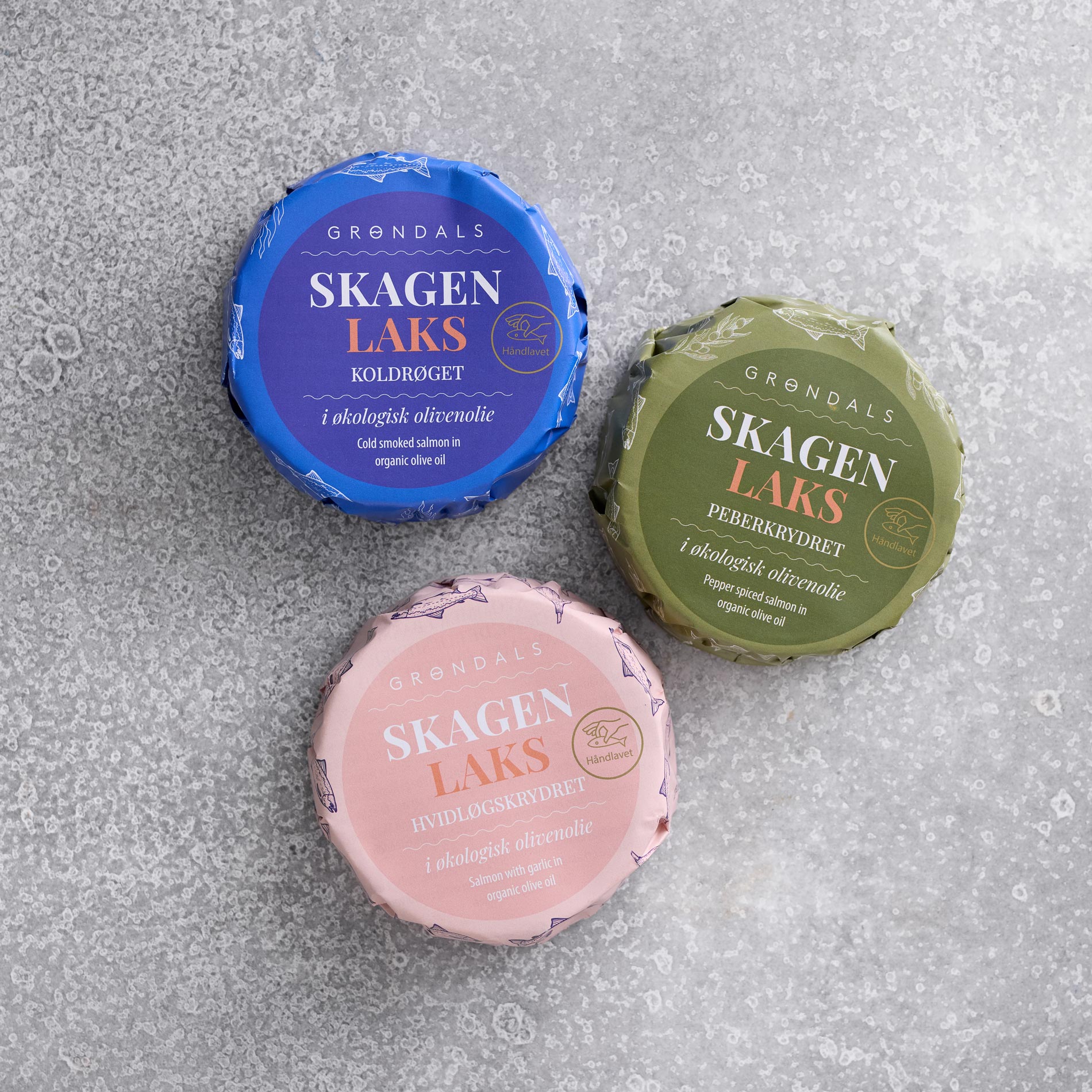 Grøndals lakse-trioen fra Grøndals omfatter tre runde dåser - koldrøget, peberkrydret og hvidløgskrydret laks - i blå, grøn og pink emballage på en grå struktureret overflade. Indeholder økologisk olivenolie.