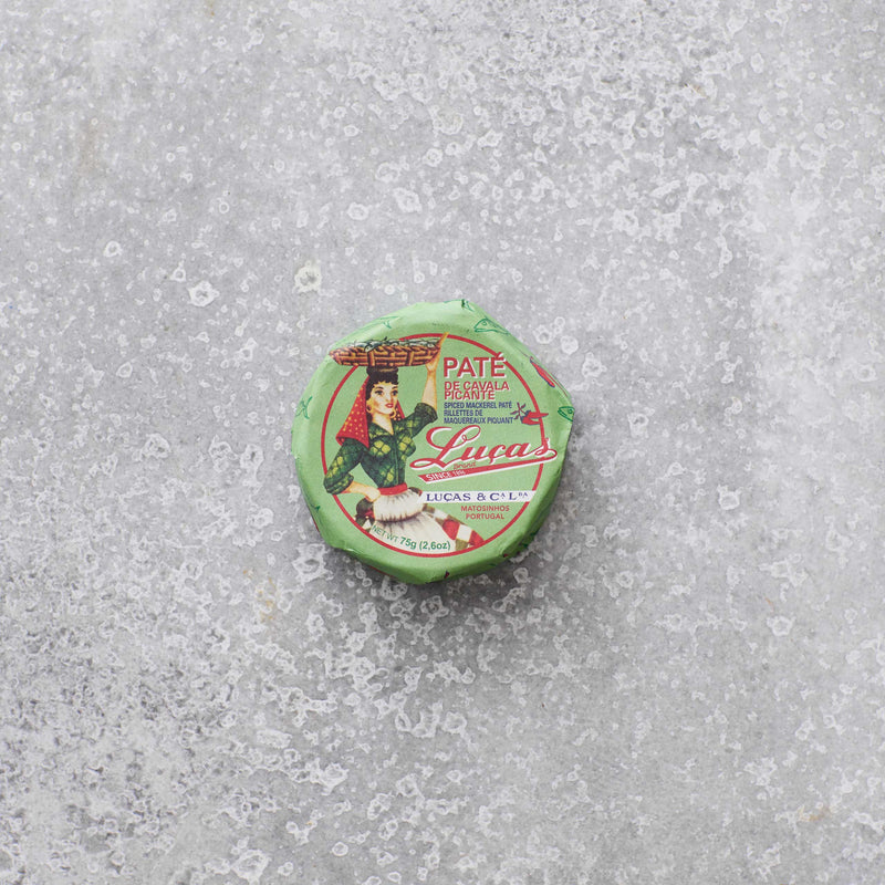 En lille, rund dåse Lucas Spicy makrelpâte med grøn emballage, rød og hvid skrift og en illustration af en person i traditionelt tøj hviler på en tekstureret grå overflade. Med et strejf af krydret tomat for ekstra smag.