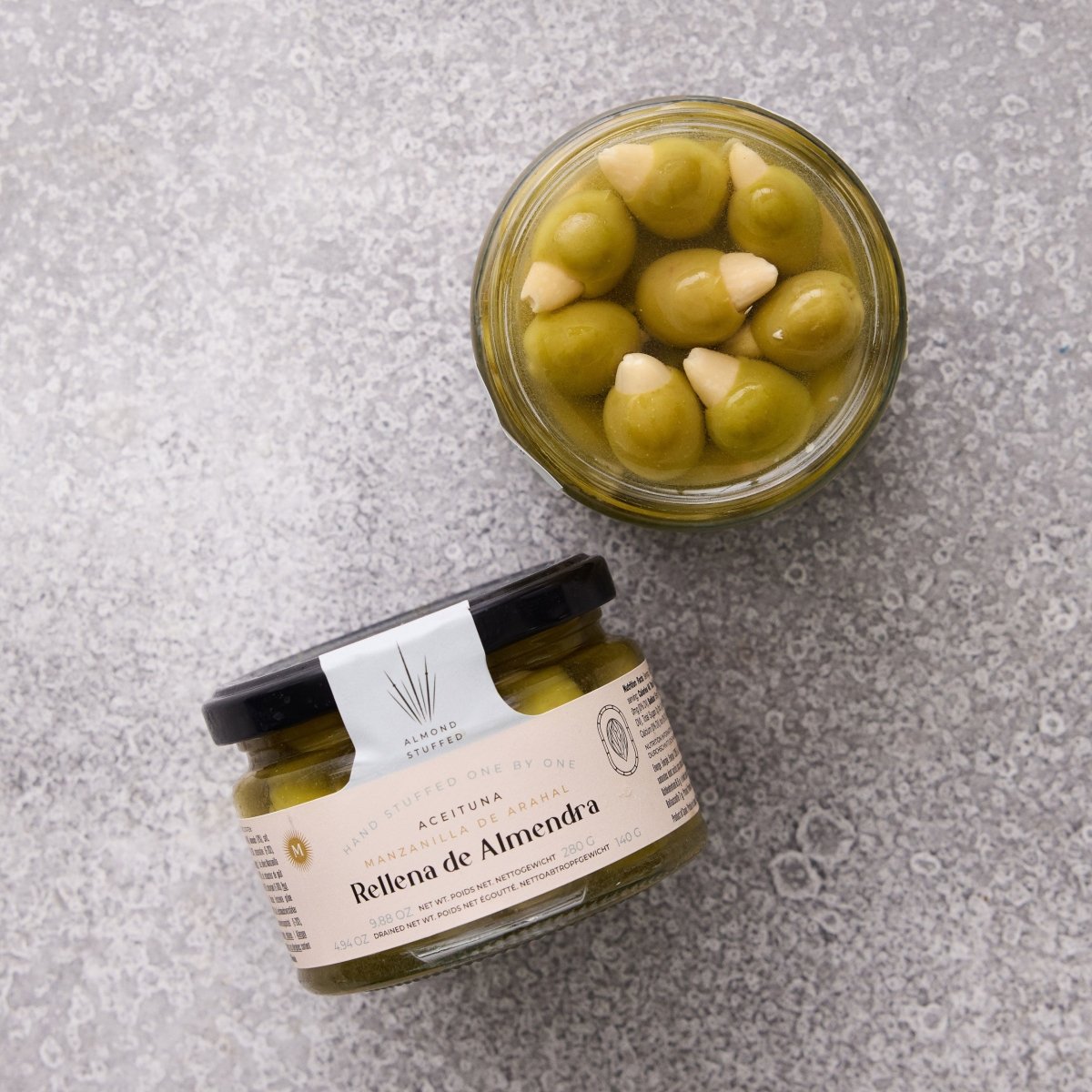 To glaskrukker med Grøndals Manzanilla oliven med mandel står på en grå stenoverflade - den ene er åben, så man kan se de mandelfyldte oliven indeni, som er ideelle til spansk tapas. Den forseglede krukke har etiketten "Rellena de Almendra.