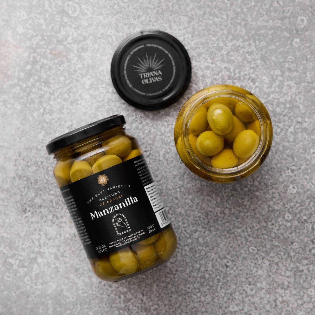 To glaskrukker med Grøndals Manzanilla oliven med sten - den ene stående med en sort etiket, den anden åben med låget i nærheden - står på en struktureret grå overflade, der er ideel for fans af spanske oliven.