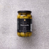 En glaskrukke med Grøndals Manzanilla oliven med sten, med sort låg og etiket, står på en struktureret grå overflade.