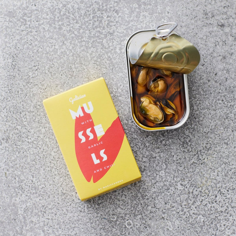 En gul æske med Don Gastronom-muslinger i chili og hvidløgsolie står ved siden af en åben dåse med marinerede muslinger i sauce og olivenolie på en grå, struktureret overflade.