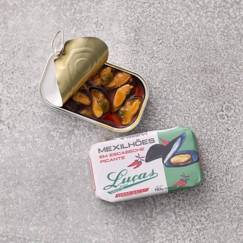 En åbnet dåse Muslinger i krydret escabeche-sauce fra Lucas ligger ved siden af emballagen, som viser en illustration af muslinger og Lucas-mærket på en grå tekstureret overflade.