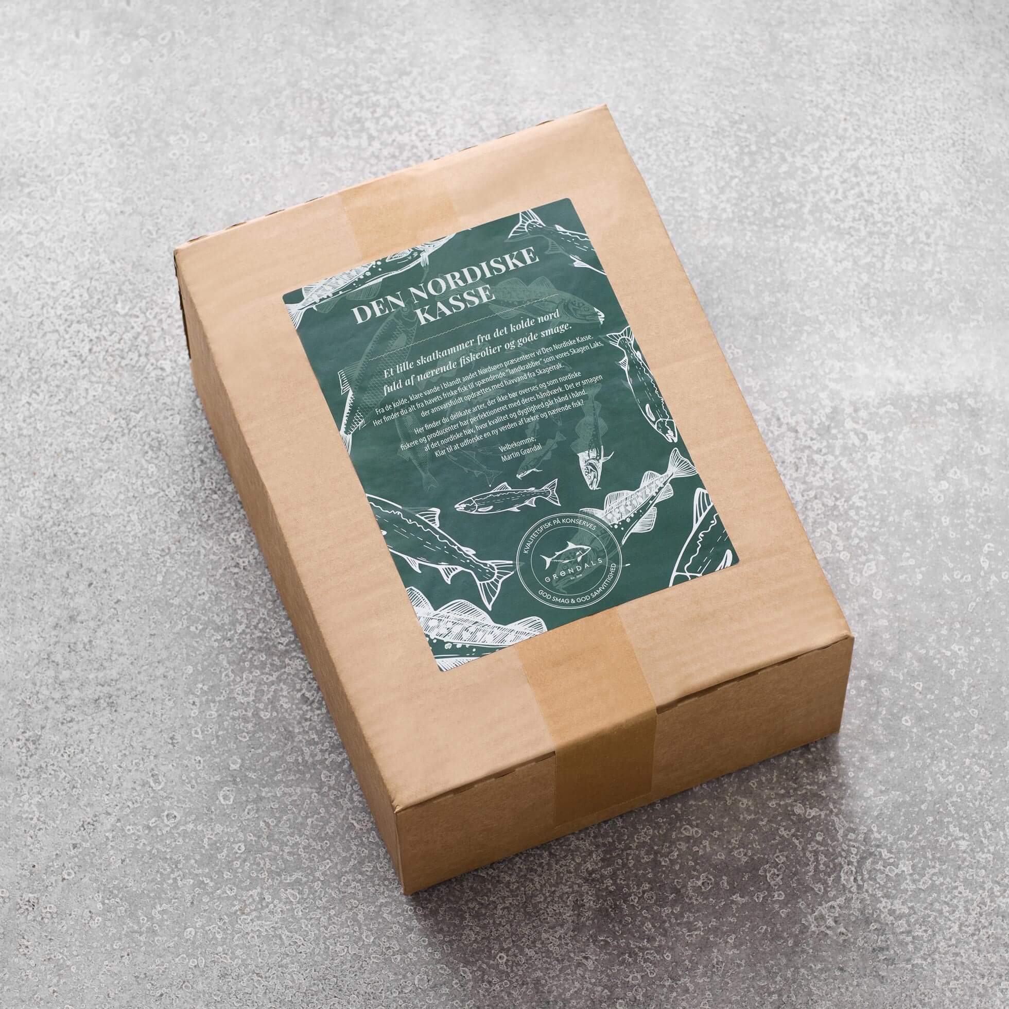 En rektangulær papkasse forseglet med brun tape står på en grå overflade. Den har en grøn og hvid etiket med titlen Den Nordiske Kasse by Grøndals, med illustrationer af nordiske fisk og dansk tekst.