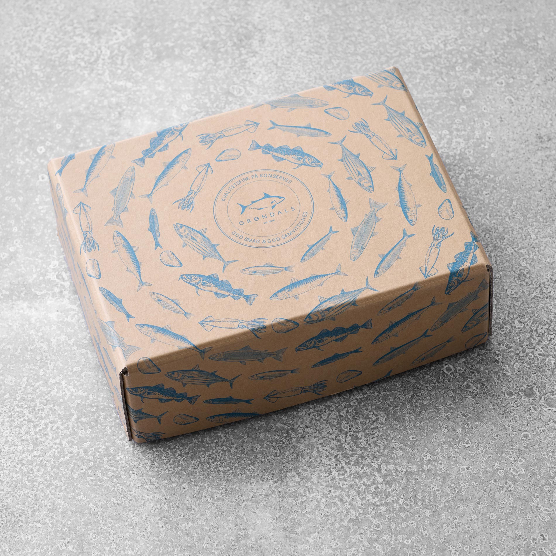 En papkasse med blå fiskeillustrationer, placeret på en tekstureret grå overflade. Grøndals smagekasse med olivenolie-æsken har et centralt cirkulært logo med tekst og fiskegrafik, der repræsenterer Grøndals-mærket.
