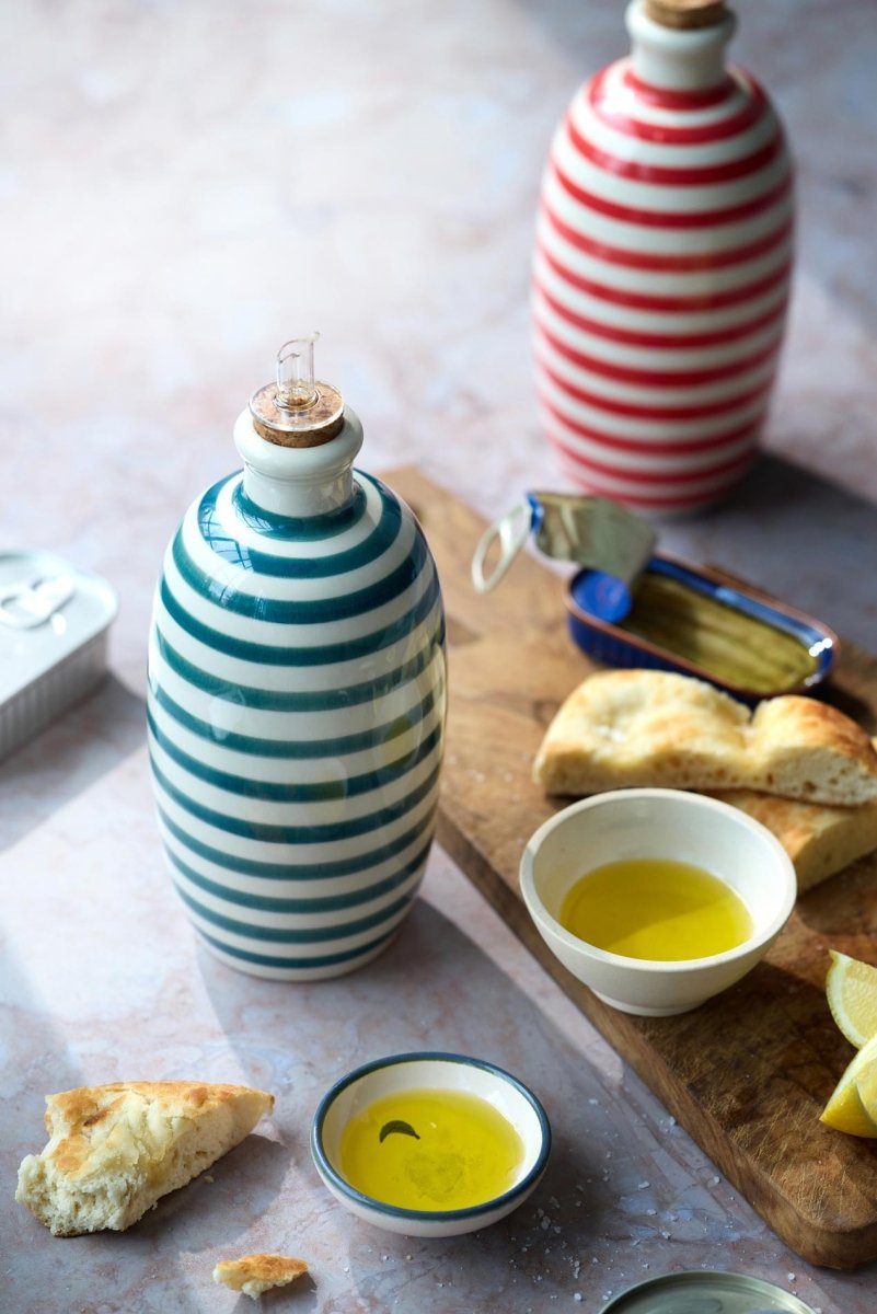 En grøn- og hvidstribet keramikflaske står ved siden af en lille skål med Grøndals Øko ekstrajomfru-olivenolie Arbequina-Picual (EVOO), brød og et træbræt. En rød- og hvidstribet flaske og mere brød ses i baggrunden.