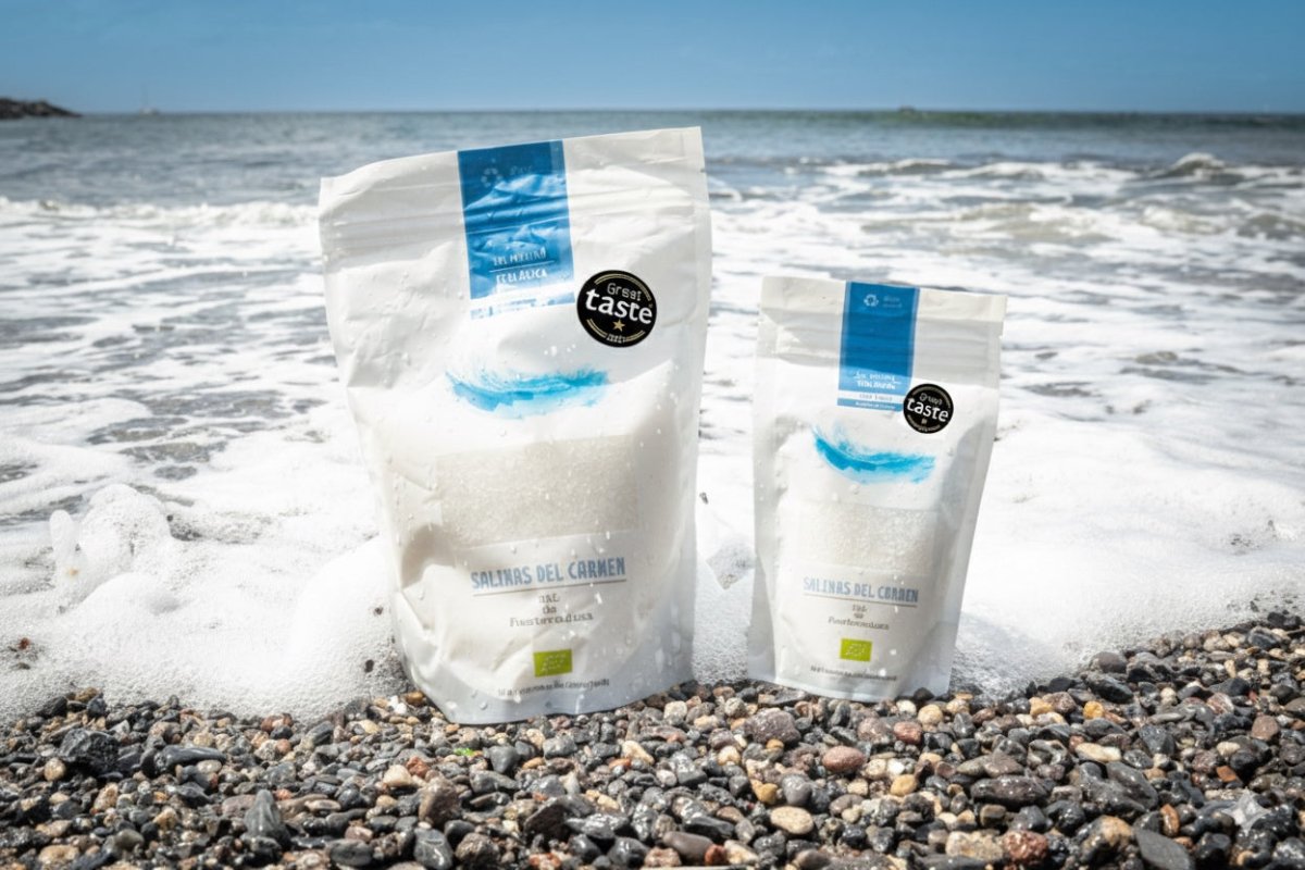 To hvide poser med »Sal de Fuerteventura Økologisk Havsalt – Håndhøstet fra Fuerteventura« ligger på en stenet strand ved havet under en blå himmel. Begge poser er forsynet med sorte »Great Taste Award«-klistermærker.