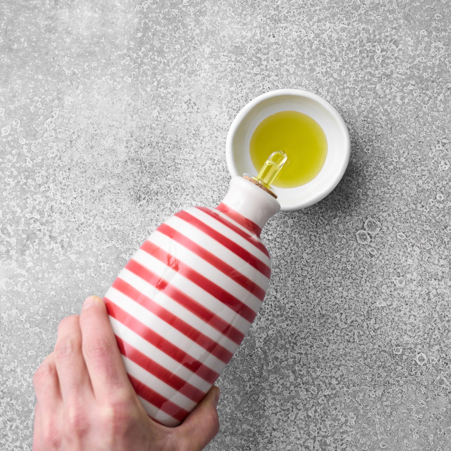 En hånd hælder Grøndals Øko ekstrajomfru-olivenolie Arbequina-Picual (EVOO) fra en keramikflaske med røde og hvide striber ned i en lille hvid skål på en grå tekstureret overflade.