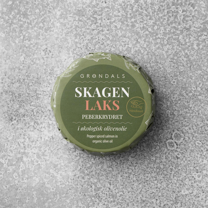 En rund dåse med grønt låg mærket Grøndals står på en gråmønstret overflade. På etiketten står der, at den indeholder Peberkrydret laks i økologisk olivenolie.