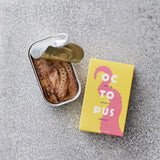 En åbnet dåse Don Gastronom Pulpo i hvidløgsolie står ved siden af en gul og lyserød æske mærket OCTOPUS på en grå tekstureret overflade.