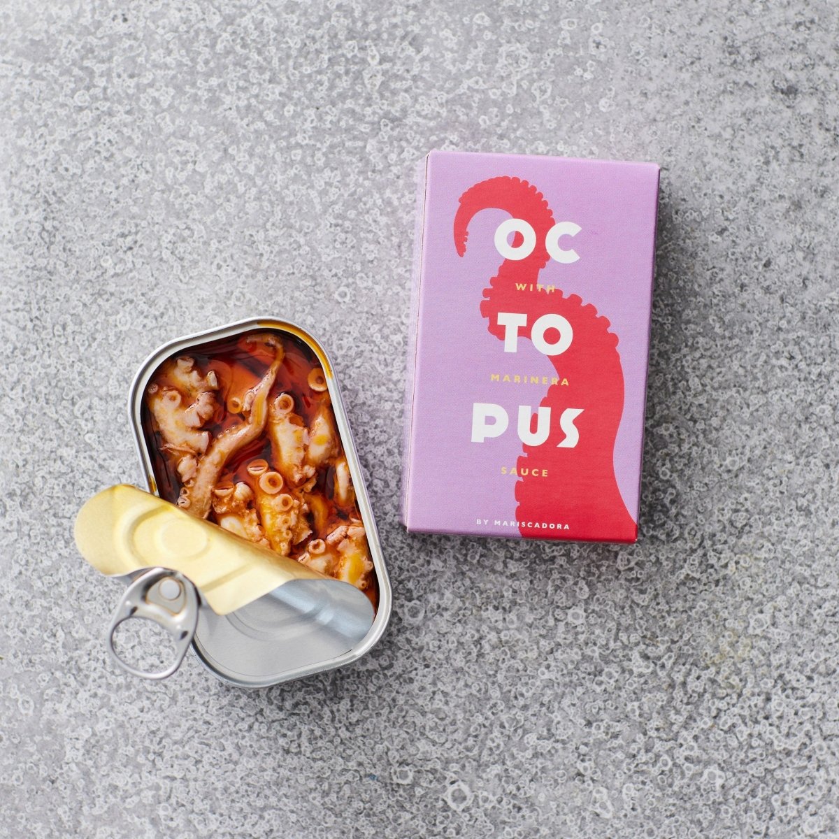 En åbnet dåse Don Gastronom Pulpo i SAUCE MARINIÈRE ligger ved siden af den livlige pink og lilla emballage med produktnavnet på en grå, struktureret overflade.