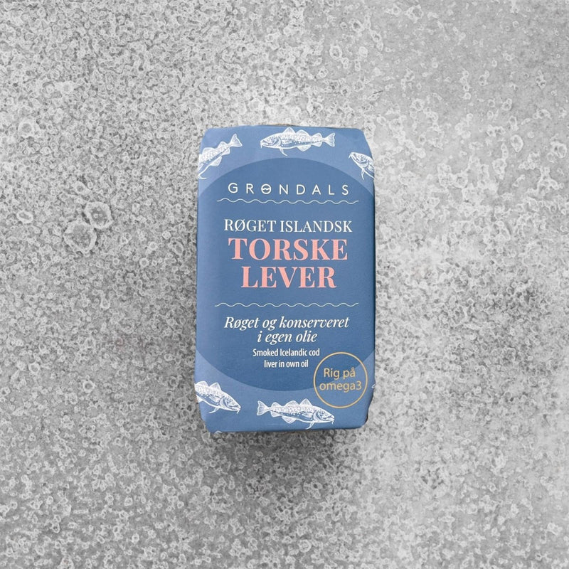 En blå dåse med Grøndals Røget islandsk torskelever i olie står på en struktureret grå overflade med hvide fiskeillustrationer og dansk tekst, der fremhæver det høje indhold af omega-3-fedtsyrer.