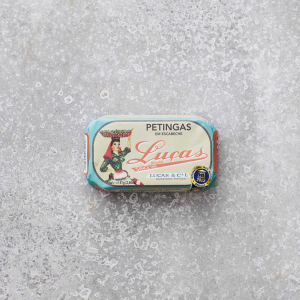 Dåse med Lucas Petingas (små sardiner) i escabeche fra Grøndals, med en illustreret etiket af en kvinde, der bærer en kurv, set mod en grå tekstureret baggrund.