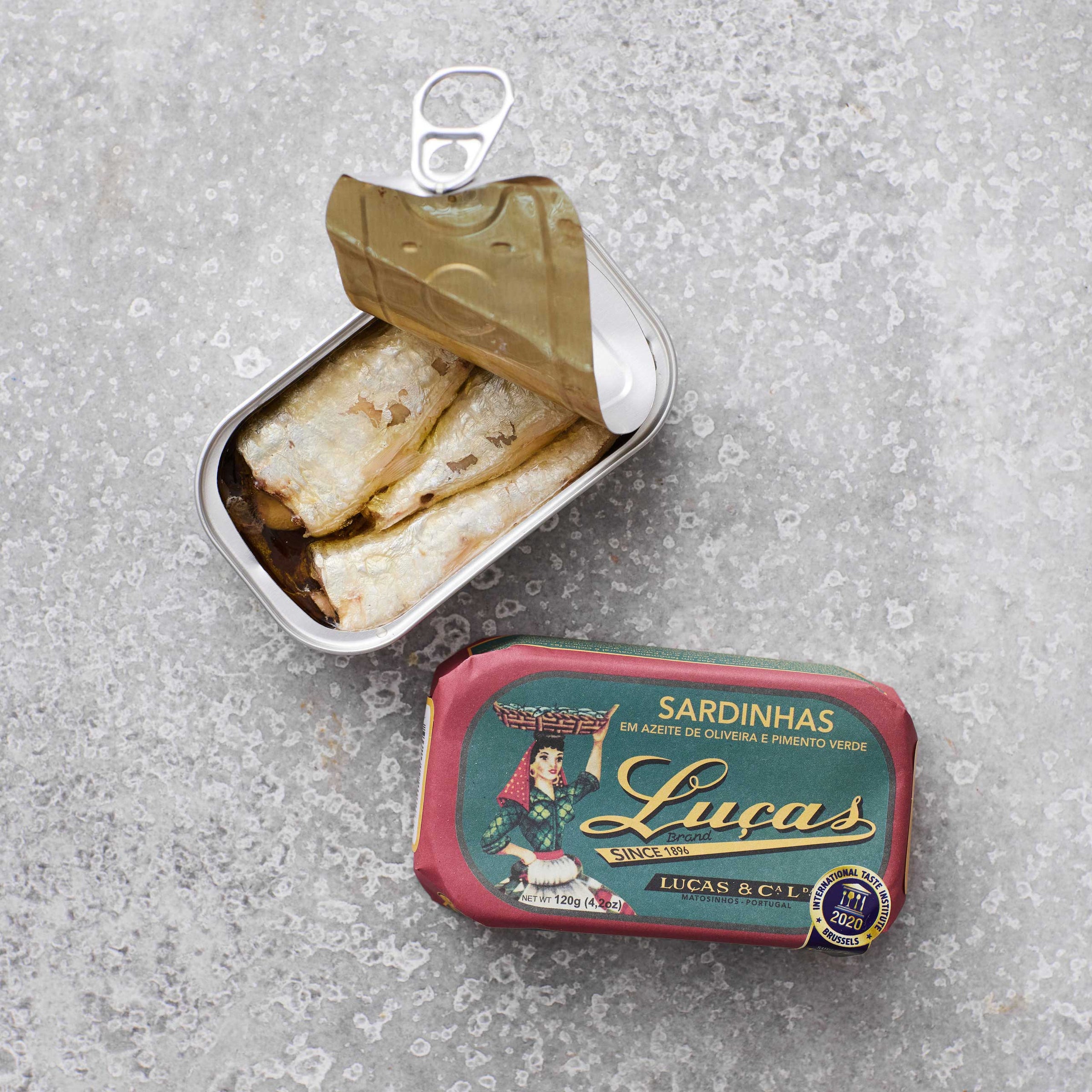 Dåse med Lucas sardiner i olivenolie og grøn peber fra Grøndals, hvor låget er delvist åbnet, placeret ved siden af emballagen, set mod en grå tekstureret baggrund.
