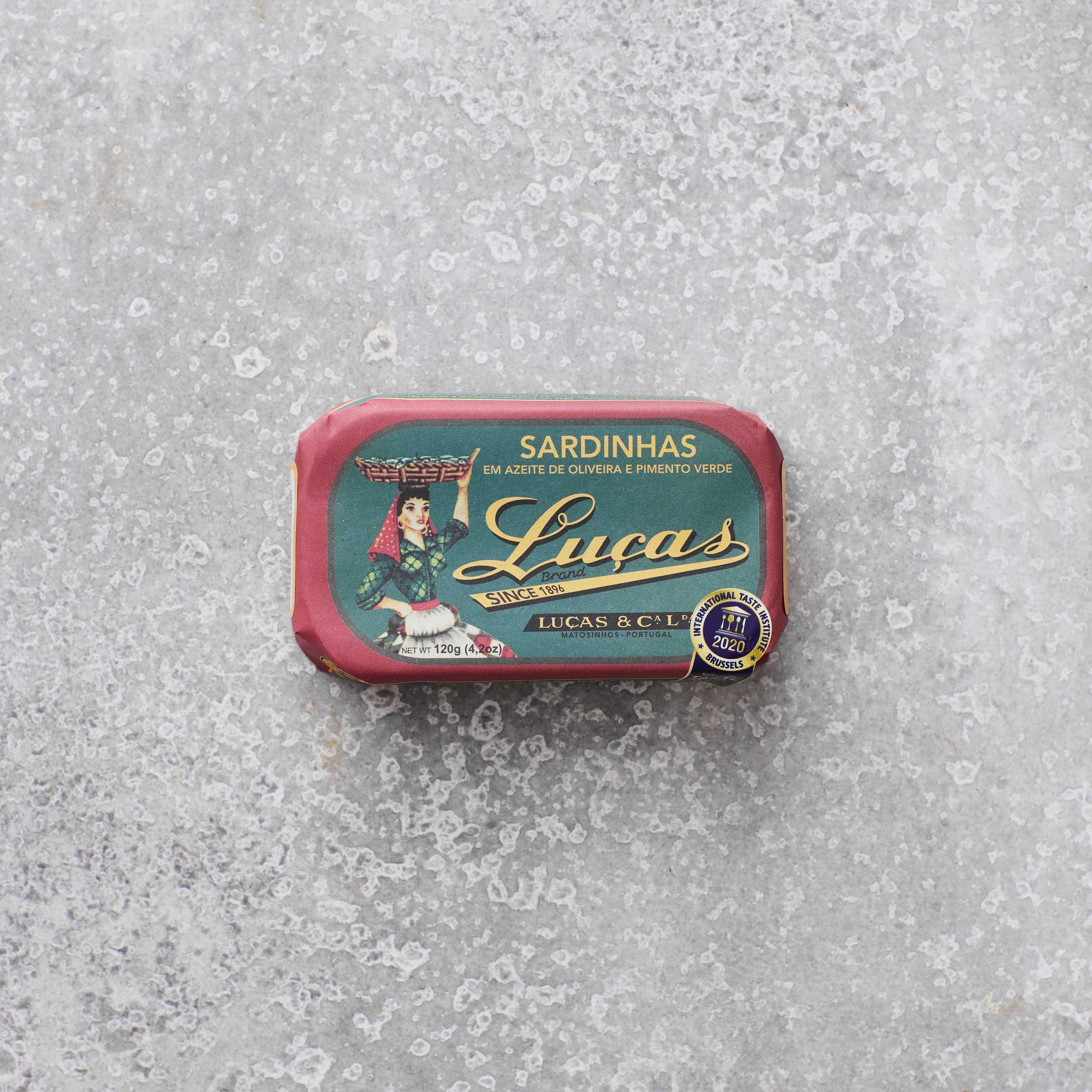 Dåse med Lucas sardiner i olivenolie og grøn peber fra Grøndals, med en illustreret etiket af en kvinde, der bærer en kurv, set mod en grå tekstureret baggrund.