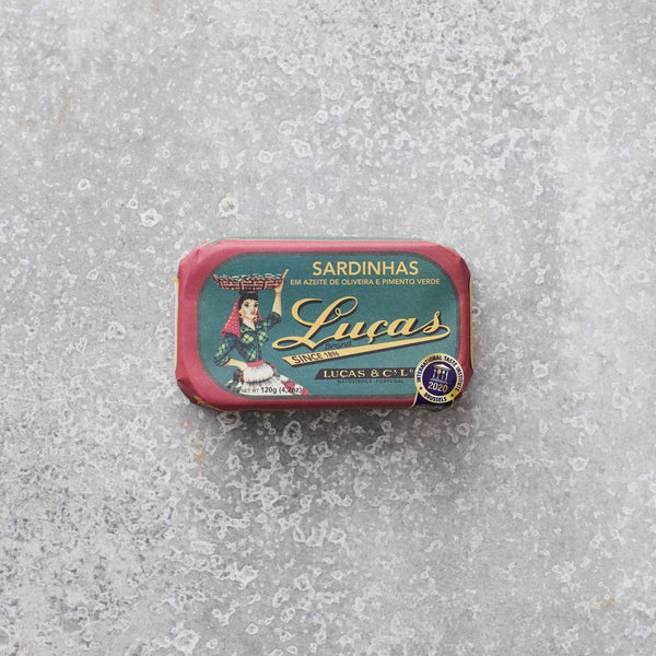 Dåse med Lucas sardiner i olivenolie og grøn peber fra Grøndals, med en illustreret etiket af en kvinde, der bærer en kurv, set mod en grå tekstureret baggrund.
