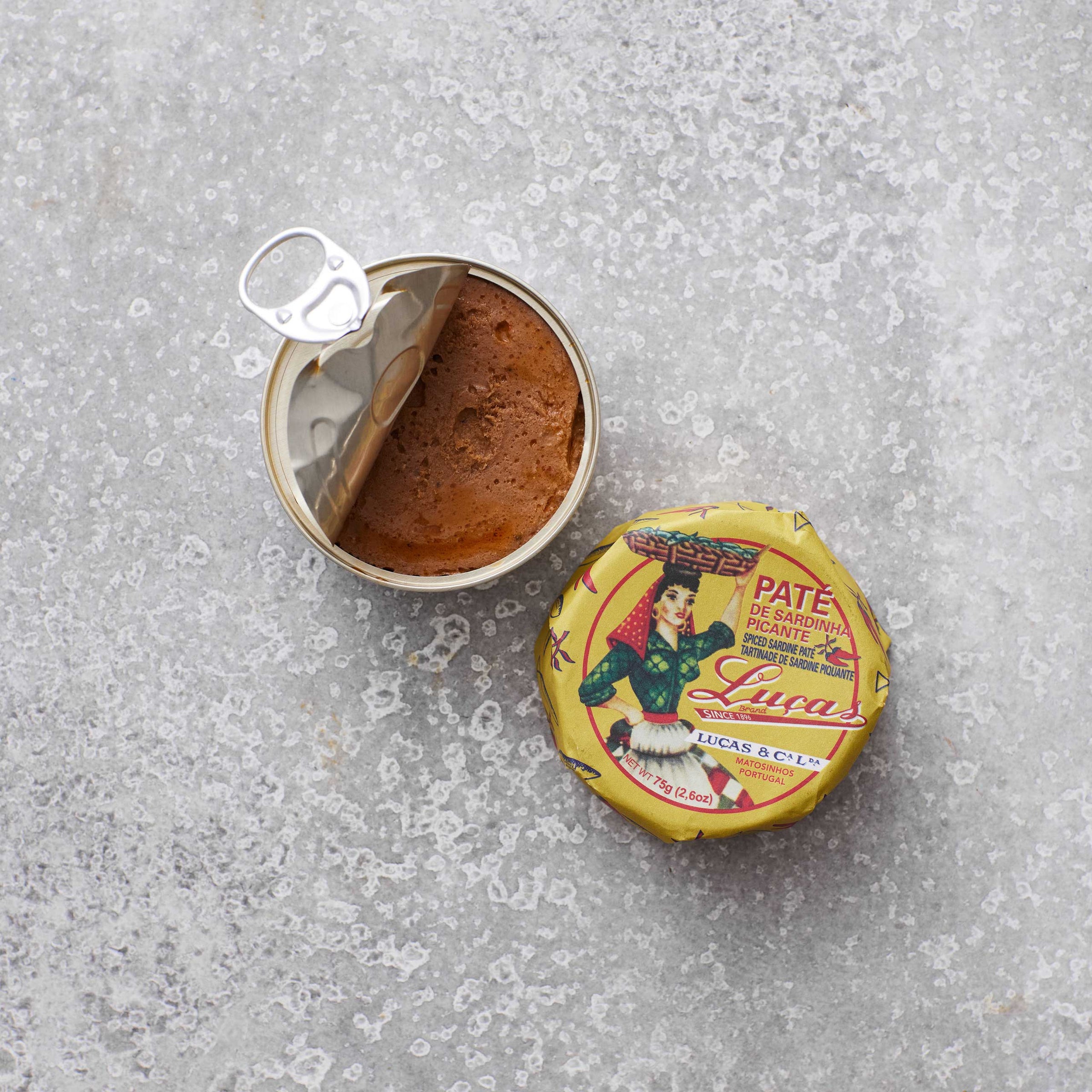 En åben dåse Lucas Spicy sardinpaté med strejf af tomat med et delvist afskrællet låg står ved siden af det farverige låg – dekoreret med en kvinde i traditionel dragt – på en lysegrå tekstureret overflade, perfekt til at smøre på friskbagt surdejsbrød.
