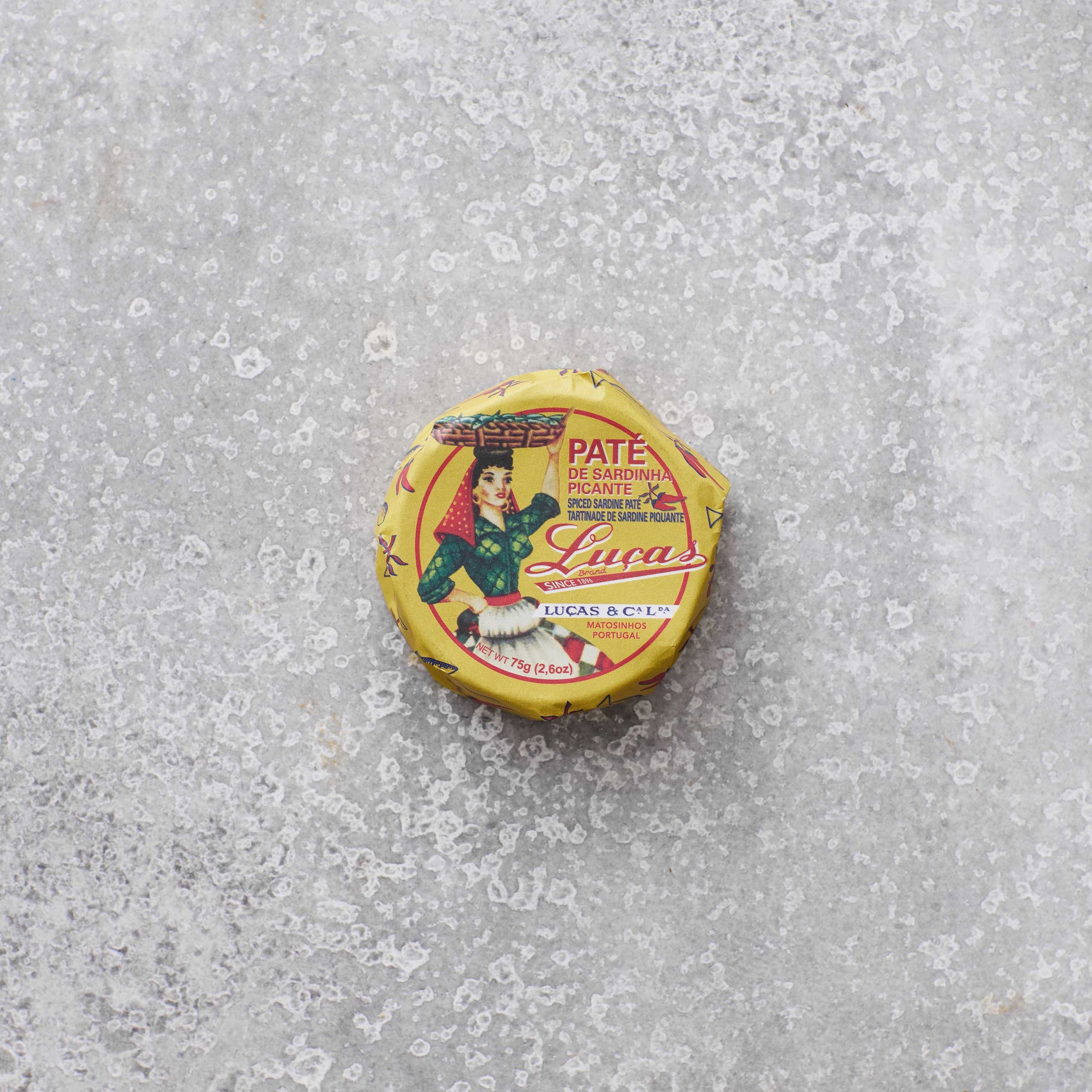 En lille rund dåse Lucas Spicy sardinpaté med strejf af tomat, hvis gule etiket viser en farverig kvinde i traditionelt klædedragt, ligger på en tekstureret grå overflade.