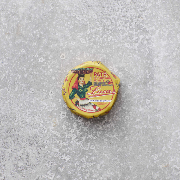 En lille rund dåse Lucas Spicy sardinpaté med strejf af tomat, hvis gule etiket viser en farverig kvinde i traditionelt klædedragt, ligger på en tekstureret grå overflade.