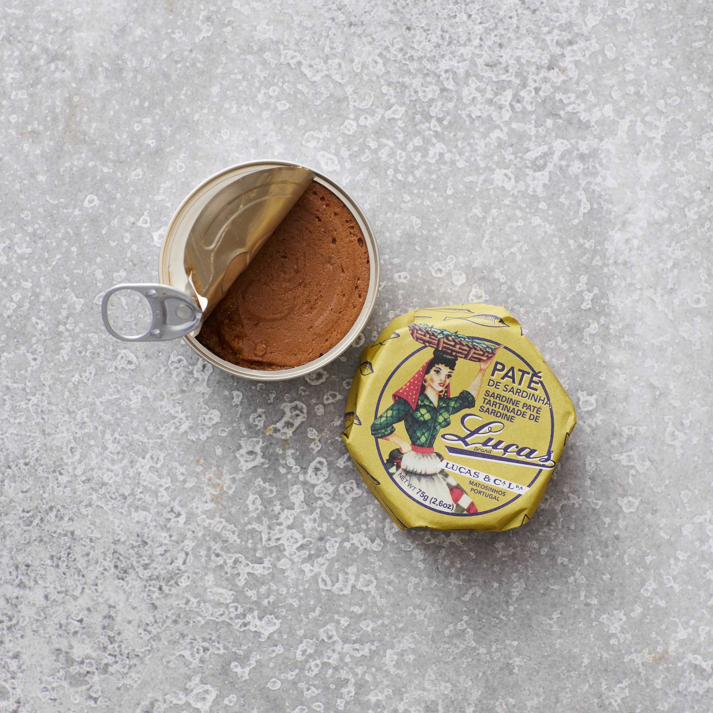 En åbnet dåse Lucas Sardinpaté med olivenolie hviler på en grå tekstureret overflade, det gyldne låg er delvist trukket tilbage. Ved siden af ligger det farverige låg med en kvinde i traditionel dragt, der antyder en fyldig olivenoliesmag.