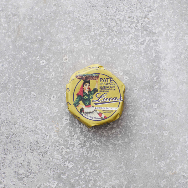 En rund, gul dåse Lucas Sardinpaté med olivenolie står på en tekstureret grå overflade, og låget har en illustration af en kvinde i traditionel klædedragt.