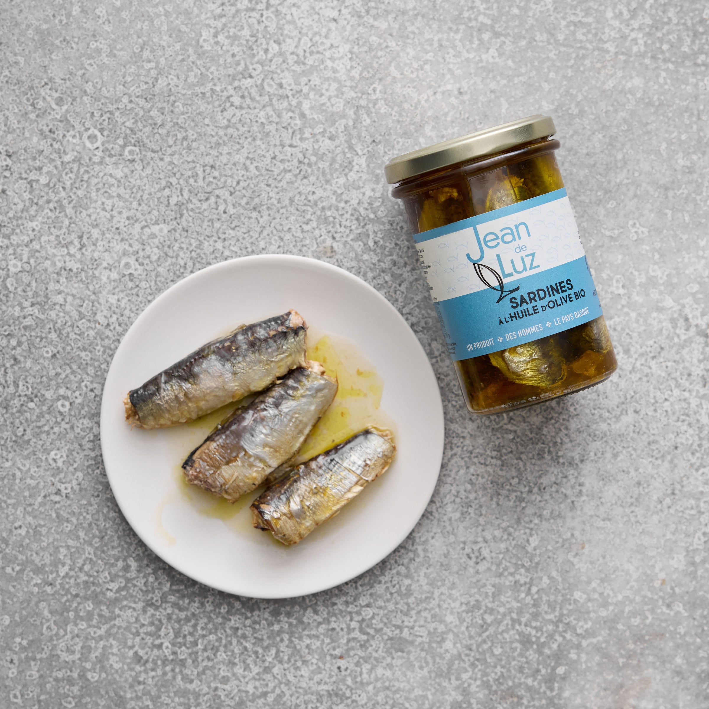 En tallerken med fire Sardiner i økologisk ekstra jomfru olivenolie står ved siden af en krukke med navnet Jean de Luz, der viser lækre økologiske sardiner på en struktureret grå overflade.