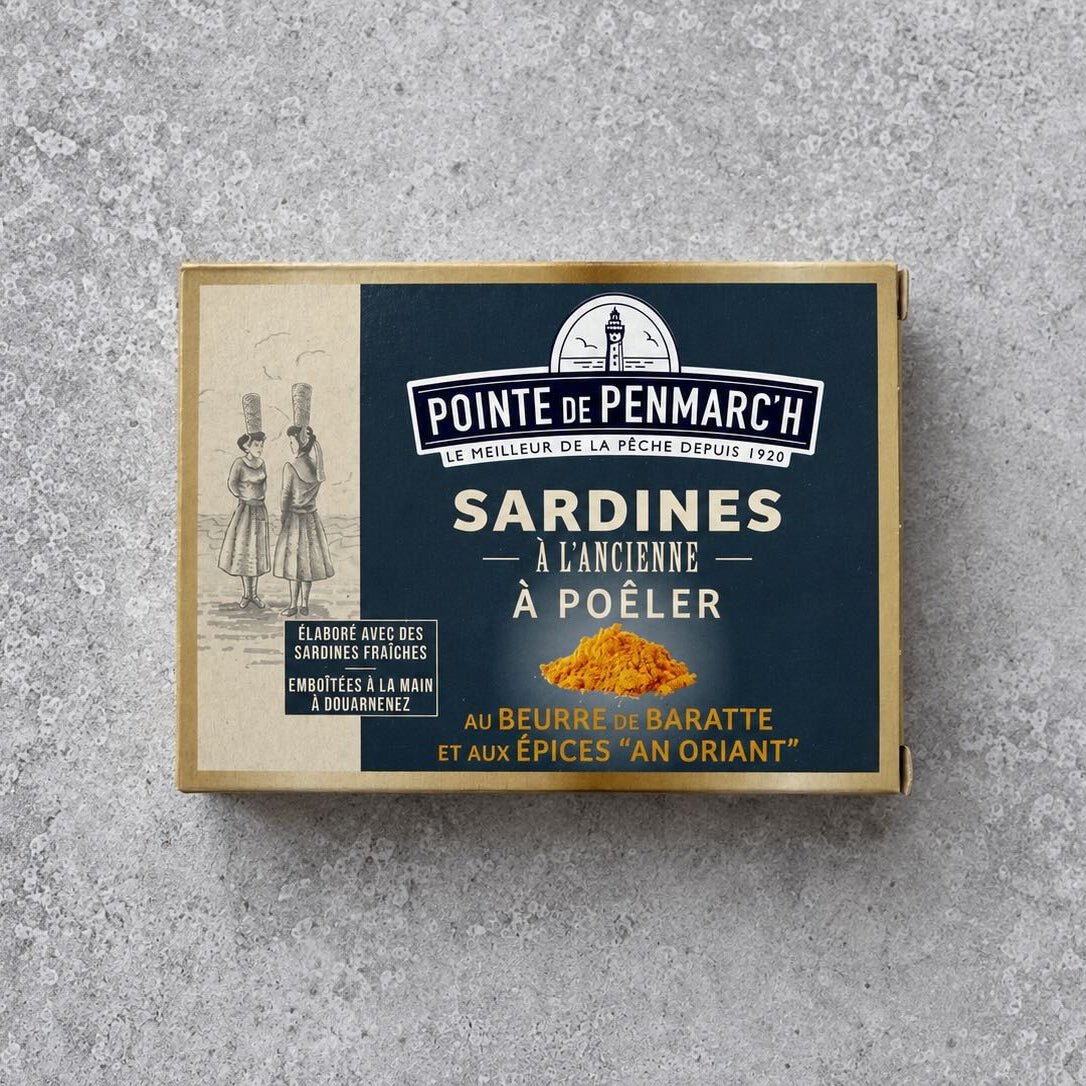 Rektangulær dåse med Pointe de Penmarc’h Sardiner med kærnet smør og karrykrydderier med en marineblå etiket, der viser kvinder med kurve, et fyrtårnslogo og en bunke gult karrypulver.