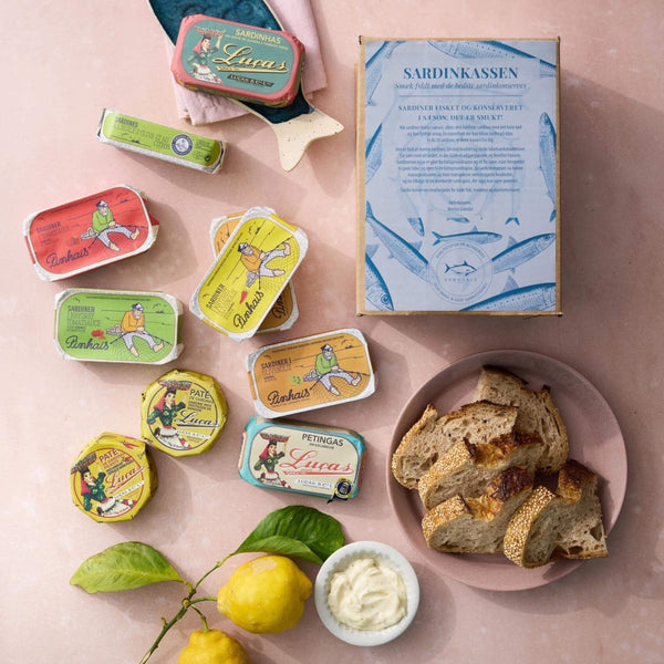 Grøndals Sardinkassen (en æske med assorterede farverige sardinpålæg), skiver brød, superfood-pålæg og to citroner med blade er arrangeret på en lyserød overflade.