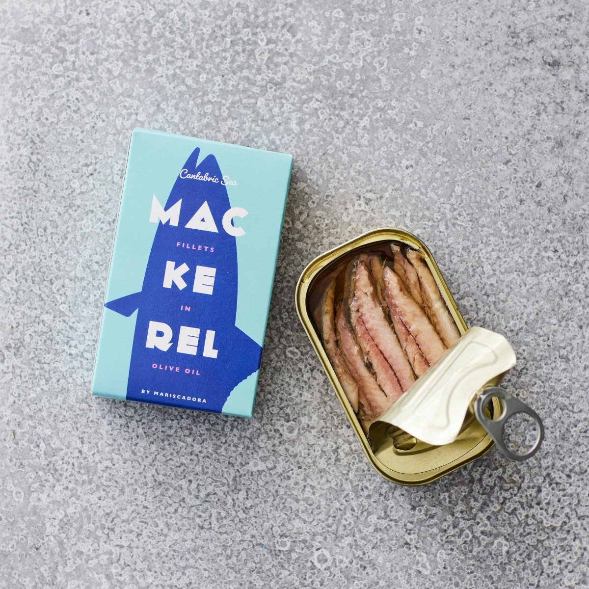 En blå æske mærket Don Gastronom Skindfri makrelfilet i olivenolie står ved siden af en åbnet dåse på en grå, struktureret overflade. Fileterne, der betragtes som superfood, er sirligt arrangeret inde i dåsen.