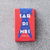 En rektangulær rød dåse med blå accenter mærket Små spanske sardiner i olivenolie af Don Gastronom er placeret på en gråplettet overflade.