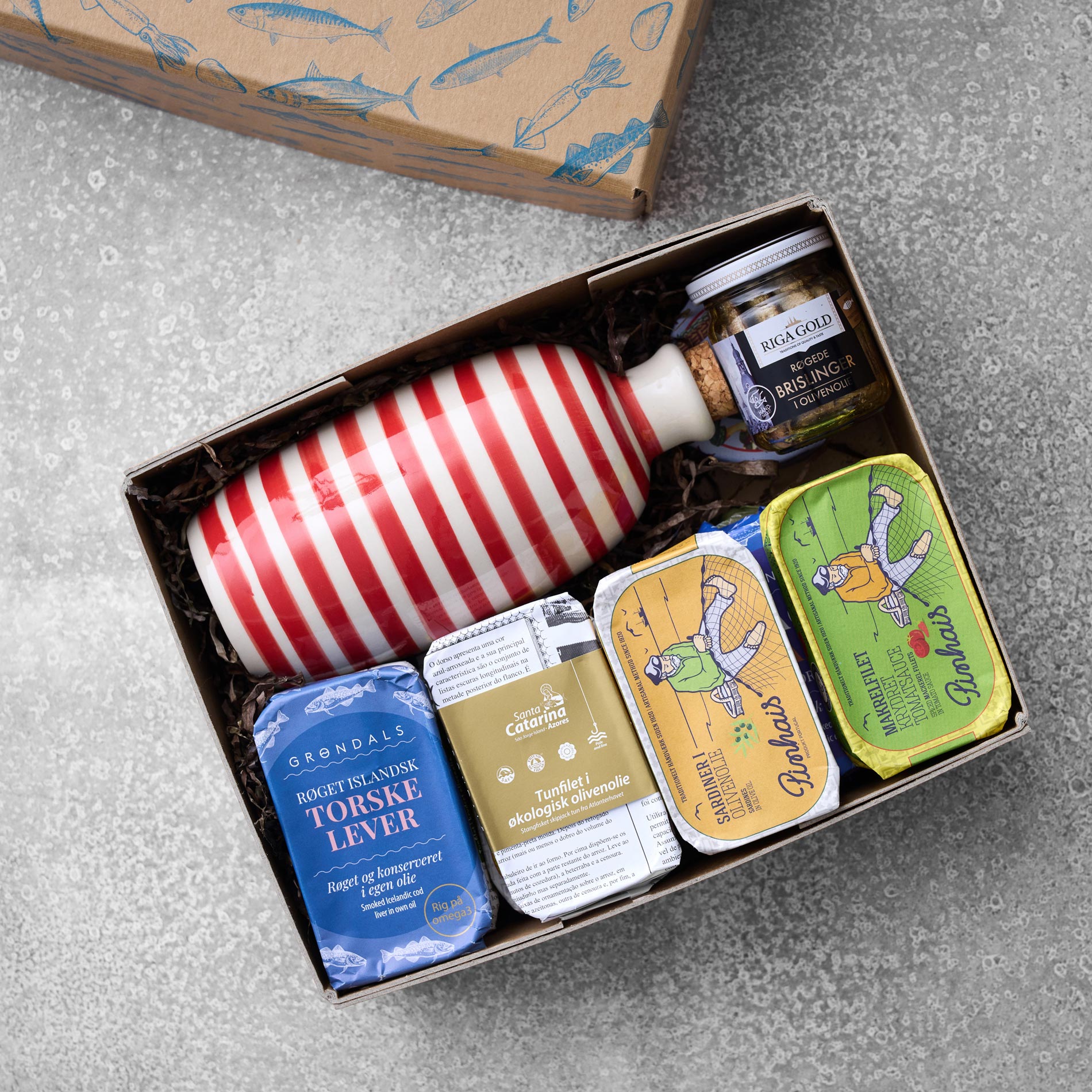 Gaveæsken Grøndals smagekasse med olivenolie fra Grøndals indeholder en stribet flaske, fire dåser fiskekonserves og et krydderiglas, alt sammen arrangeret på strimlet papir i en dekorativ æske med blå fiskeillustrationer.