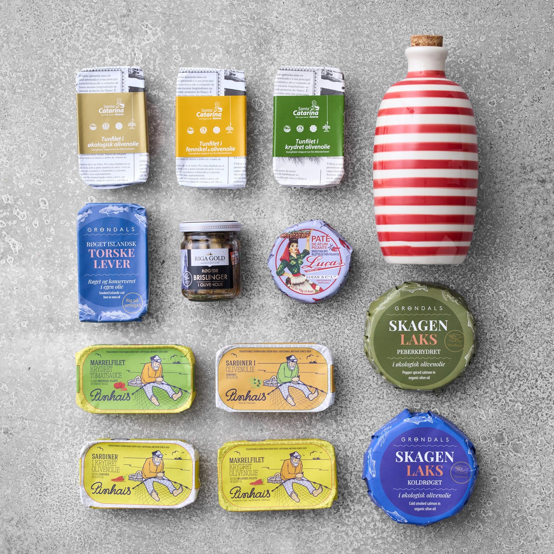 Et udvalg af Grøndals fiskekonserves, paté og krydderier fra Grøndals smagekasse med olivenolie er arrangeret på en grå overflade med farverig illustreret emballage og en rød-hvidstribet keramikflaske øverst til højre.