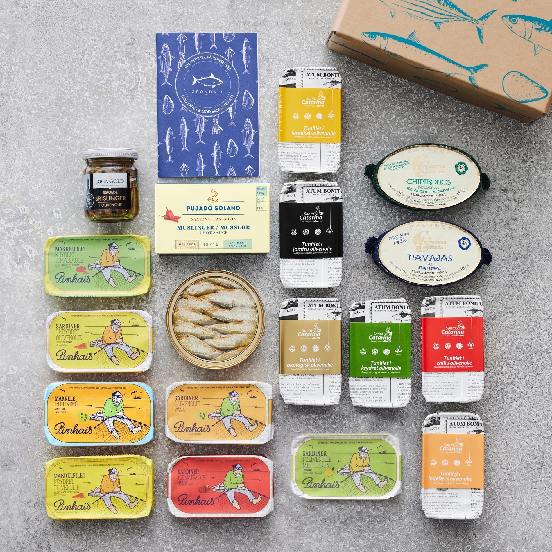 En Grøndals Smagekasse gaveæske med diverse dåser og glas med sardiner, muslinger, knivmuslinger og patéer i farverig emballage er pænt udstillet på en grå overflade.