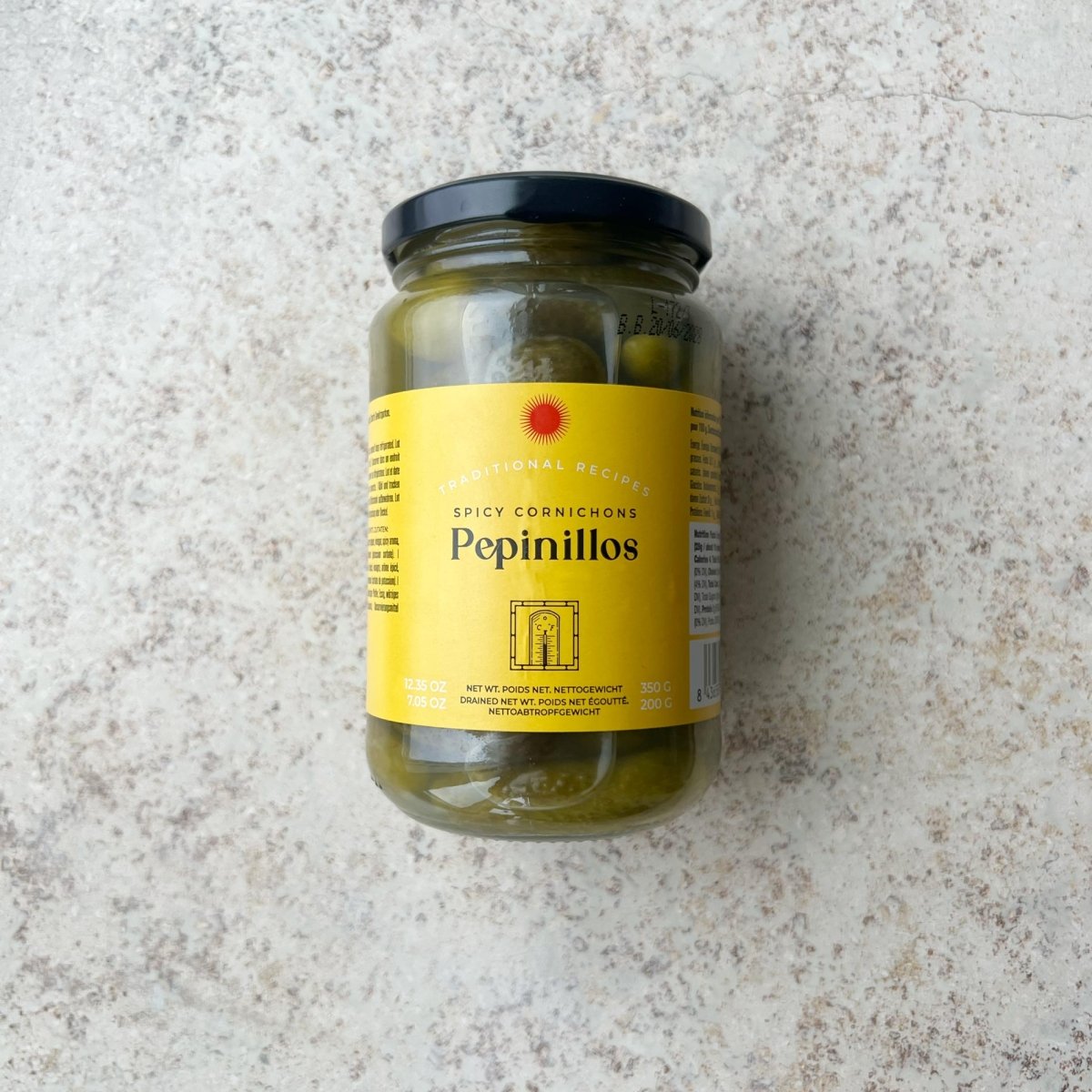 En glaskrukke med Spanske delikatesse cornichoner fra Don Gastronom med en gul etiket og sort låg er præsenteret på en lys, tekstureret overflade; perfekt til servering til tapas.