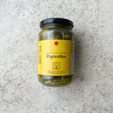 En glaskrukke med Spanske delikatesse cornichoner fra Don Gastronom med en gul etiket og sort låg er præsenteret på en lys, tekstureret overflade; perfekt til servering til tapas.