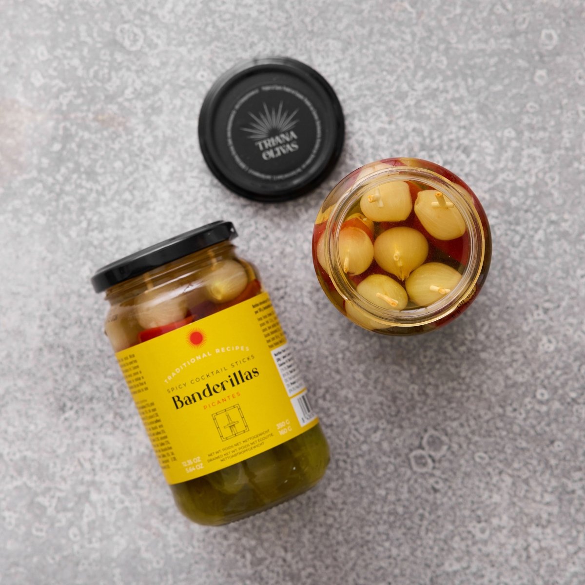 En krukke med Grøndals Spicy Banderillas cocktailpinde ligger på siden ved siden af en åben krukke med syltede løg, oliven og peberfrugter og skaber en levende snack på en grå overflade. Krukkens låg hviler i nærheden.
