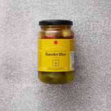Et glas med Grøndals Spicy Banderillas cocktailpinde med sort låg og gul etiket med rød tekst står på en lysegrå struktureret overflade - disse syltede grøntsager er den perfekte snack.