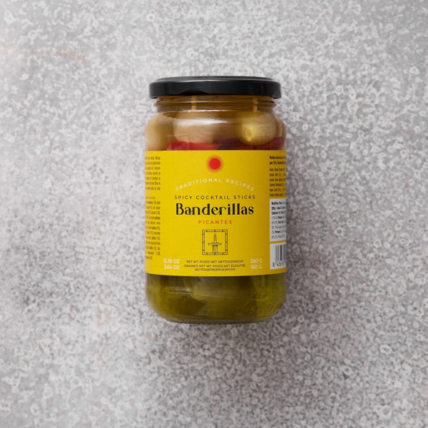 Et glas med Grøndals Spicy Banderillas cocktailpinde med sort låg og gul etiket med rød tekst står på en lysegrå struktureret overflade - disse syltede grøntsager er den perfekte snack.