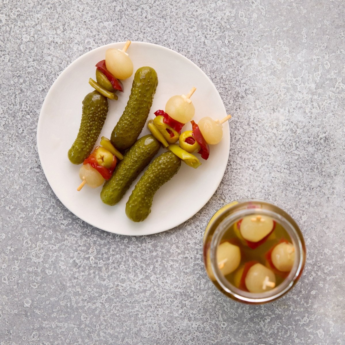 På en hvid tallerken står Grøndals Spicy Banderillas cocktailpinde - spyd af syltede oliven, peberfrugter og løg - og udgør en livlig snack. Hver krydret bid holdes sammen af en pind med et åbent glas med lignende pickles ved siden af.