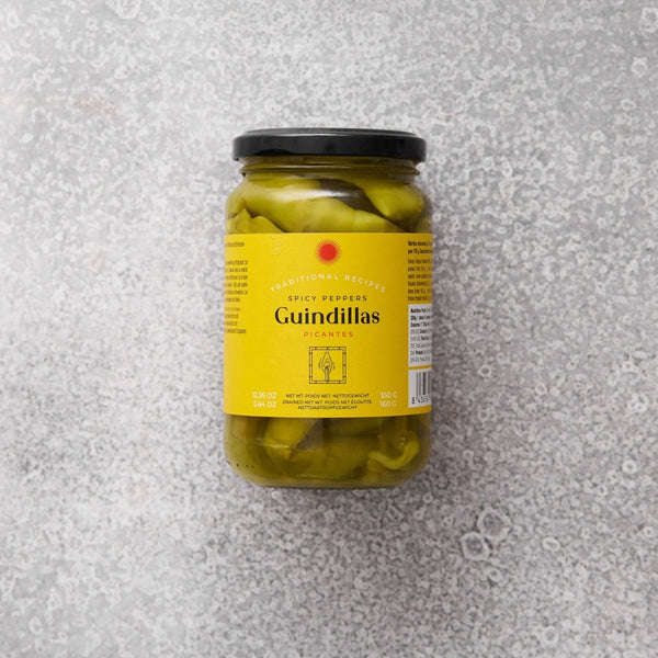 En glaskrukke med Grøndals Spicy guindillas peber med gul etiket og sort låg hviler på en struktureret grå overflade. Den tosprogede etiket antyder, at de er ideelle til tapas.
